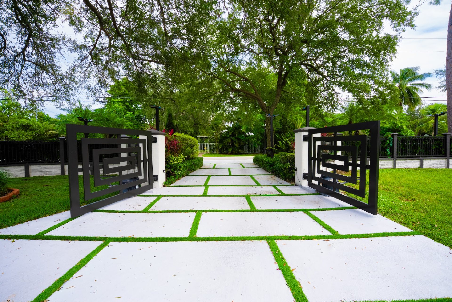Concrete driveway slabs with turf joints leading to a black geometric aluminum gate between white pillars