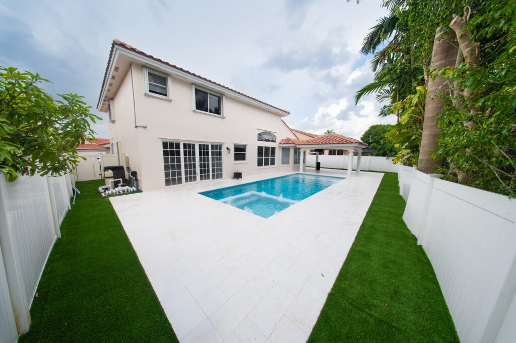Renovated pool and spa with a white deck and turf borders behind a two-story home