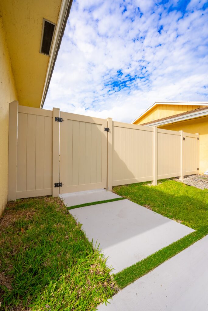 Tan vinyl privacy fence and gate beside a home with a concrete and turf walkway